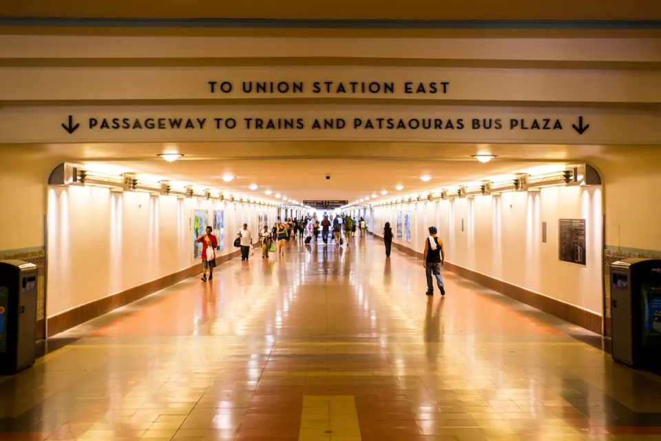 View looking down indoor concourse at Los Angeles Union Station with “To Union Station East” and “Passageway to Trains and Patsaouras Bus Plaza” signs.